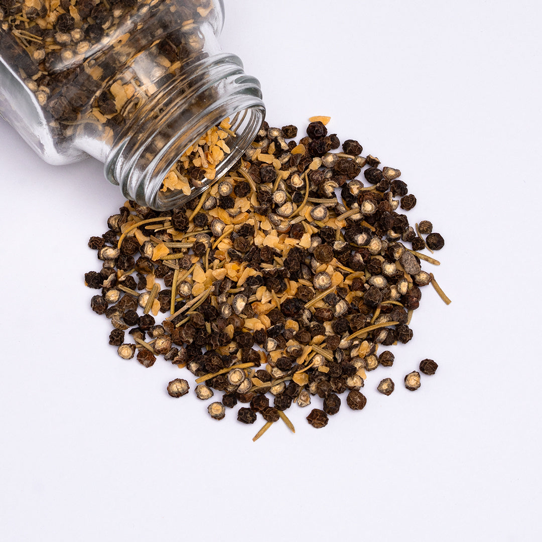 Cowgirl Peppercorn blend spilling from a glass jar, showing dried peppercorn, minced garlic, and rosemary on a white surface.
