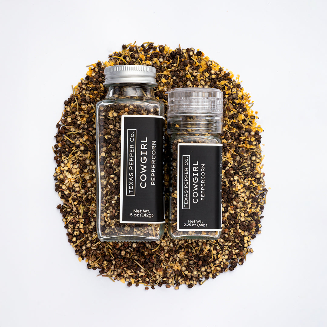 Top-down view of Texas Pepper Co. Cowgirl Peppercorn 5 oz and 2.25 oz jars placed on a bed of coarse black peppercorn, garlic, and rosemary.