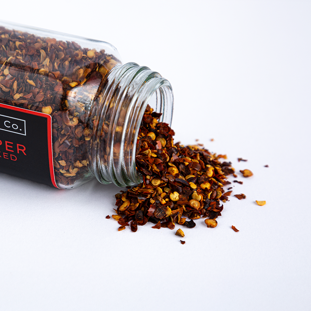 Texas Pepper Co. Crushed Red Pepper Pecan Smoked – Glass jar tipped over with smoked red pepper flakes spilling onto white surface