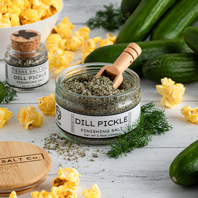 Dill pickle seasoning jar with popcorn and cucumbers on a wooden surface