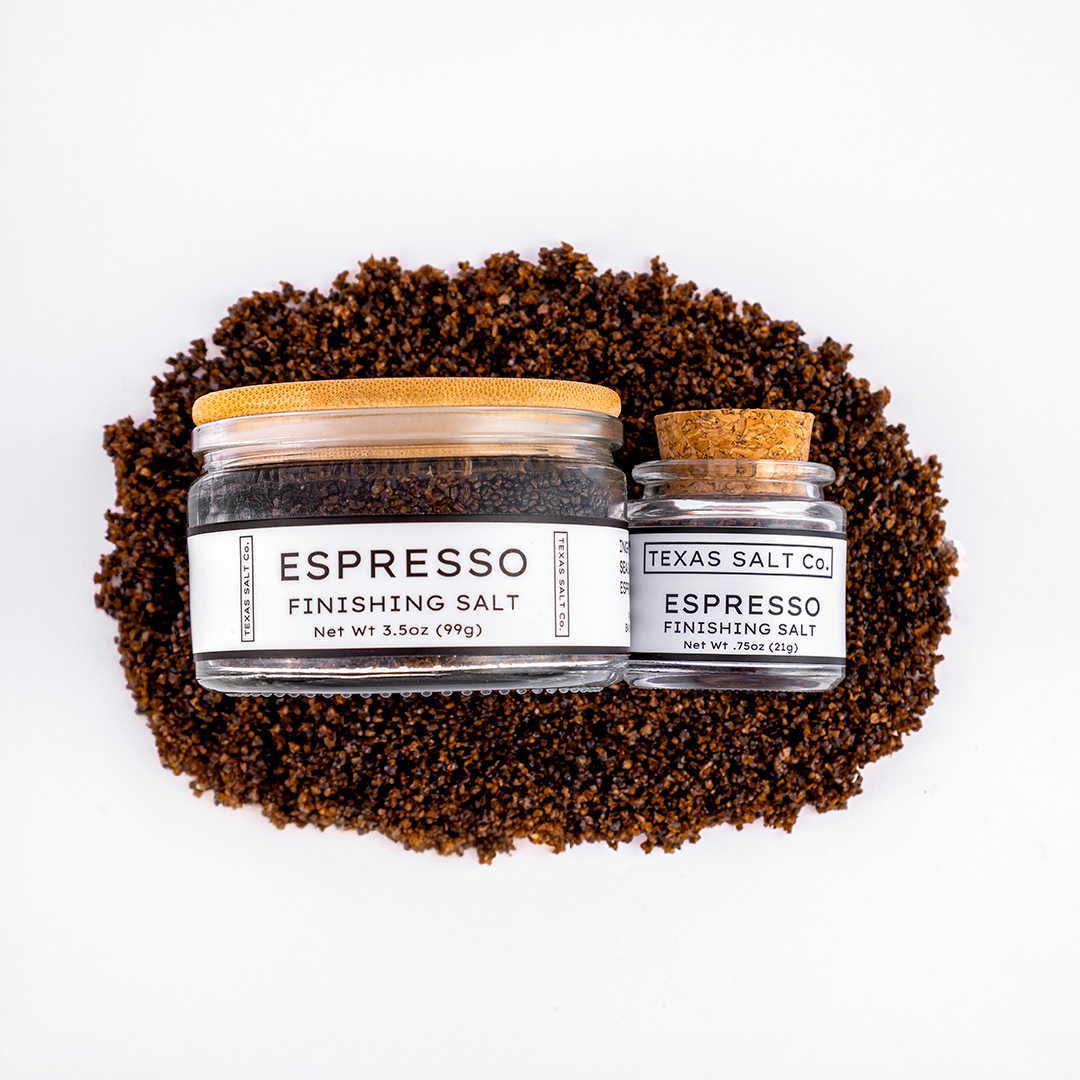 Top-down view of Texas Salt Co. Espresso Finishing Salt, showing 3.5 oz jar with bamboo lid and 0.75 oz jar with cork lid, placed on a bed of dark espresso-infused salt.