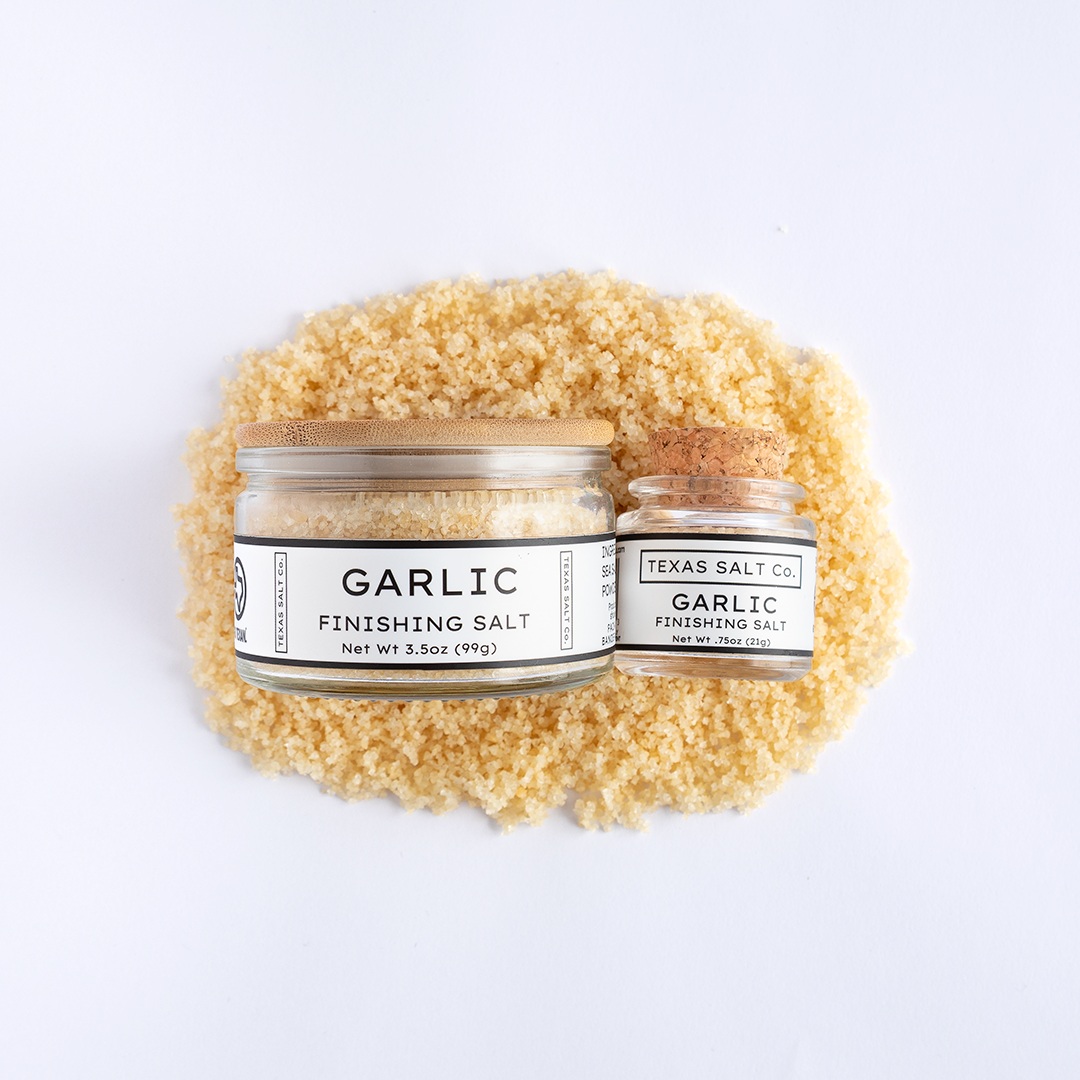 Top-down view of Texas Salt Co. Garlic Finishing Salt jars (3.5 oz and 0.75 oz) placed on a bed of garlic salt.