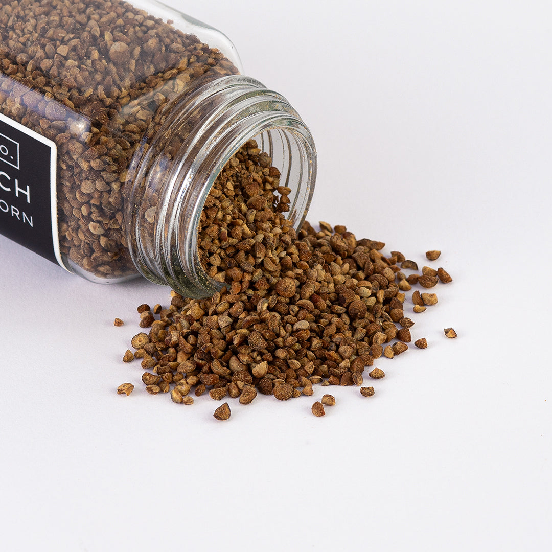 Texas Pepper Co. White Peach Smoked Peppercorn spilling out of glass jar onto white surface, showing golden smoked cracked pepper texture.