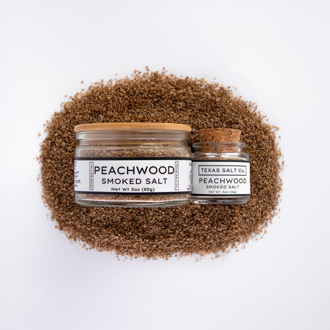 Top-down view of Texas Salt Co. Peachwood Smoked Salt in 3 oz bamboo-lid jar and 0.5 oz cork-lid jar, placed on a bed of peachwood smoked salt.