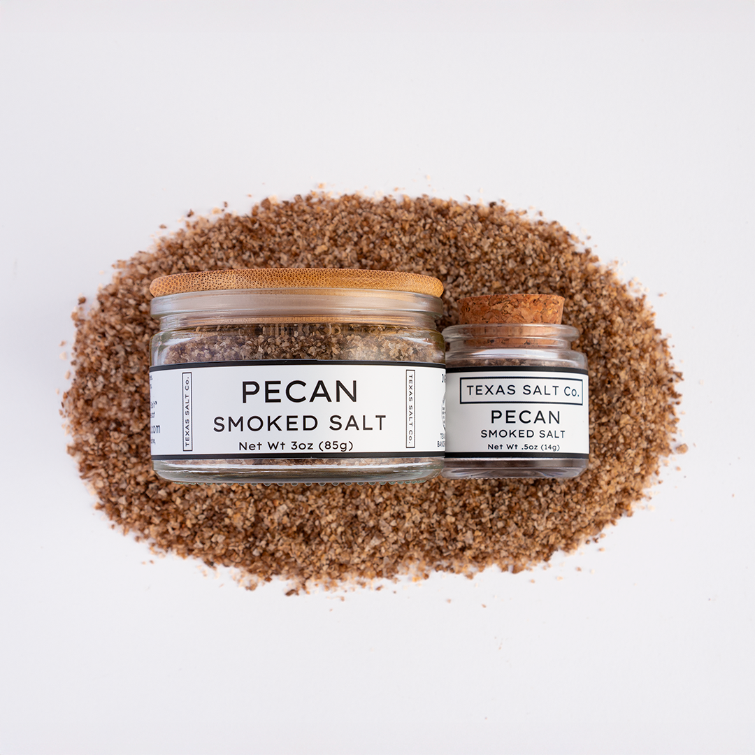 Top-down view of Texas Salt Co. Pecan Smoked Salt, showing 3 oz jar with bamboo lid and 0.5 oz jar with cork lid, placed on a bed of coarse pecan smoked salt.