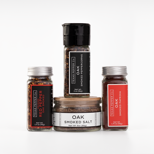 Four Texas Salt Co. products arranged together on a clean white background: a tall grinder of smoked paprika, a jar of smoked salt, a bottle of red pepper flakes, and a bottle of oak smoked paprika.
