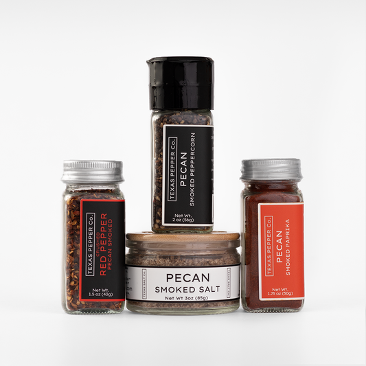 Four containers from Texas Pepper Co. arranged together on a clean white background: a tall grinder labeled pecan smoked peppercorn, a jar of pecan smoked red pepper flakes, a jar of pecan smoked paprika, and a jar of pecan smoked salt.