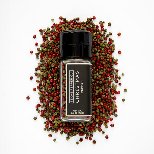 A bottle of Texas Smoked Co. Christmas Pepper surrounded by scattered red and green peppercorns on a clean white background.