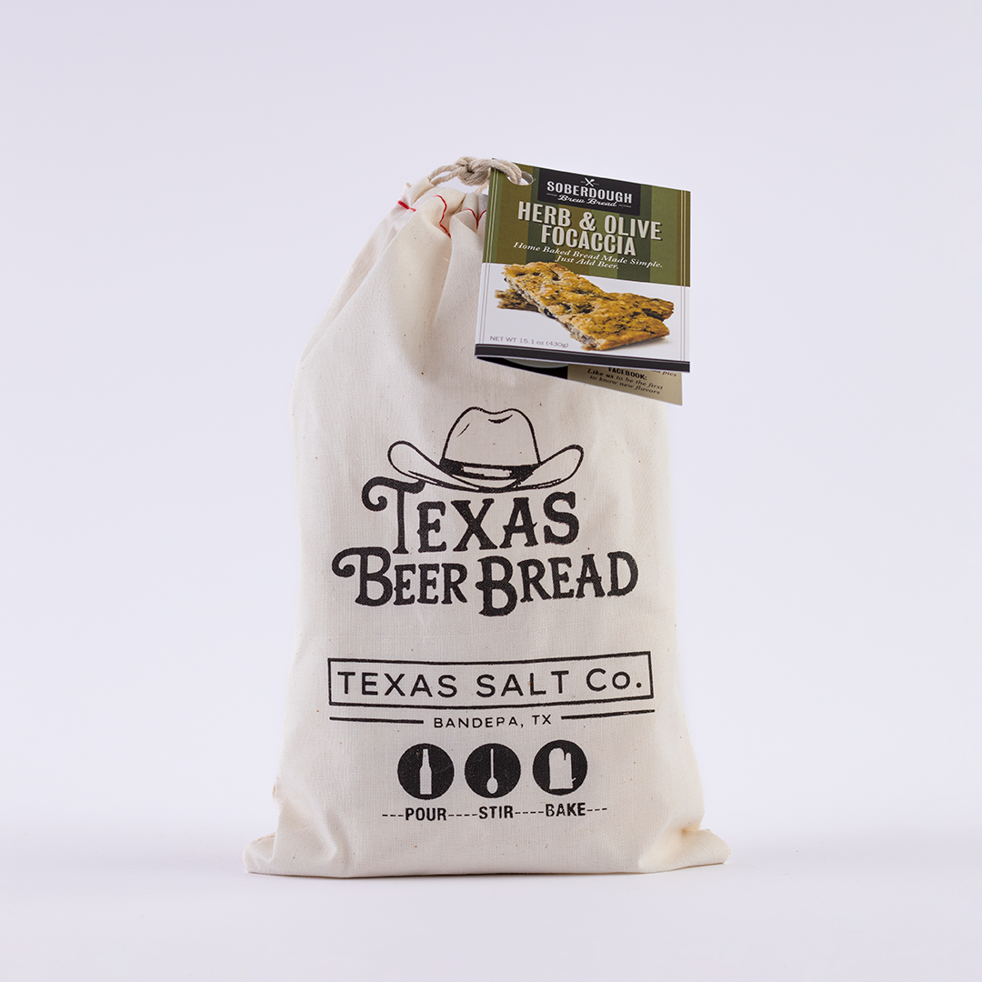 Texas Salt Co. Texas Beer Bread mix in a cloth bag with a Herb & Olive Focaccia flavor tag attached, labeled with “Pour, Stir, Bake” instructions.