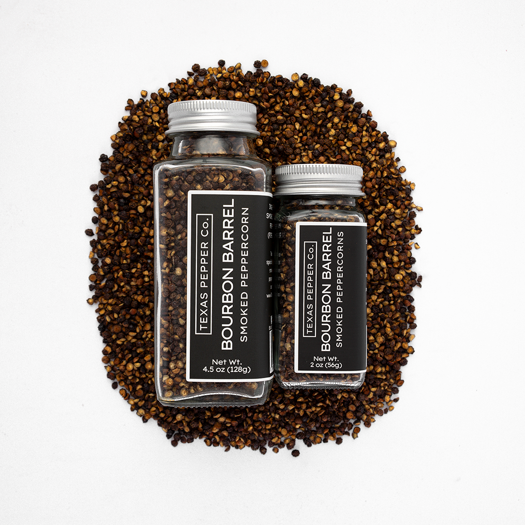 Two bottles of Texas Pepper Co. Bourbon Barrel Smoked Peppercorns surrounded by peppercorns on a white background.