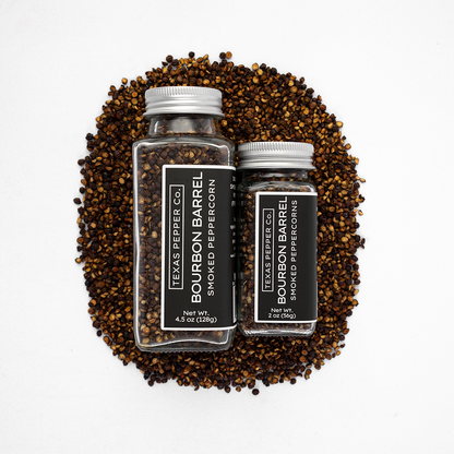 Two bottles of Texas Pepper Co. Bourbon Barrel Smoked Peppercorns surrounded by peppercorns on a white background.