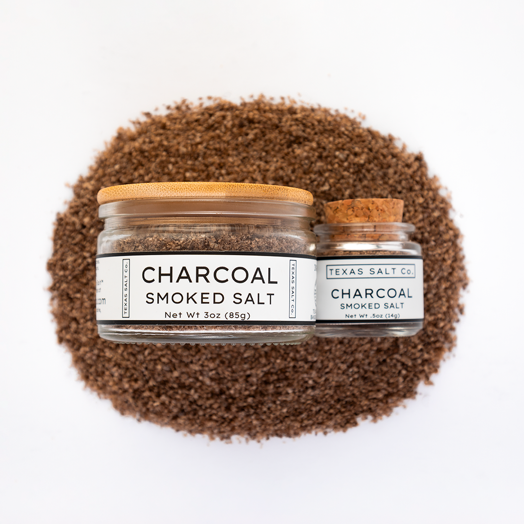Texas Salt Co. Charcoal Smoked Salt 3oz and .5oz jars displayed on top of a circular pile of charcoal-smoked salt in a top-down composition.