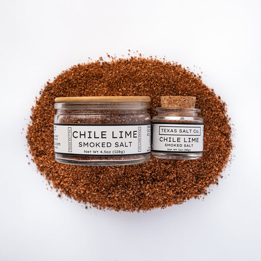 Top-down view of Texas Salt Co. Chile Lime Smoked Salt, showing 4.5 oz bamboo jar and 1 oz cork jar on a bed of reddish smoked salt.