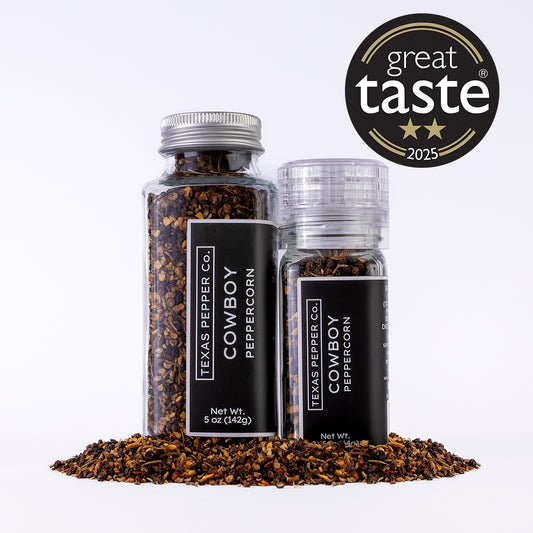 Two glass jars of Texas Pepper Co. Cowboy Peppercorn (2.25 oz grinder and 5 oz jar) with spilled smoked peppercorn blend in front.