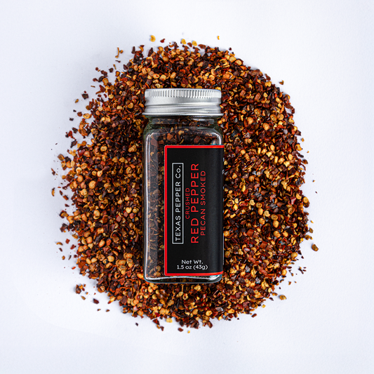 Texas Pepper Co. Crushed Red Pepper Pecan Smoked – Overhead view of glass jar on scattered bed of smoked crushed red pepper flakes