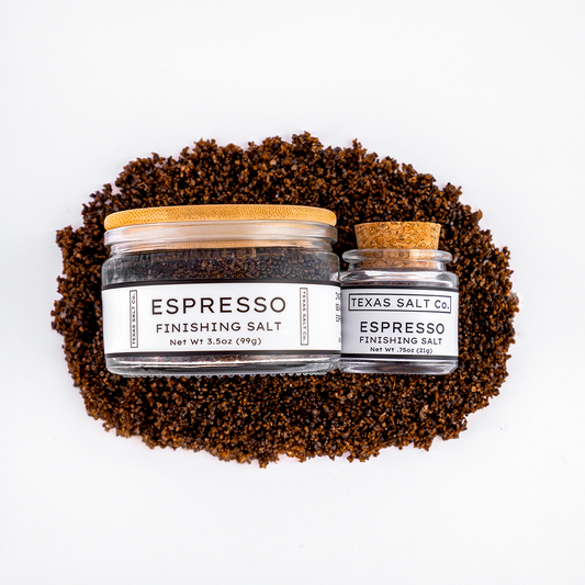Top-down view of Texas Salt Co. Espresso Finishing Salt, showing 3.5 oz jar with bamboo lid and 0.75 oz jar with cork lid, placed on a bed of dark espresso-infused salt.