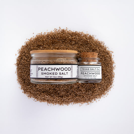 Top-down view of Texas Salt Co. Peachwood Smoked Salt in 3 oz bamboo-lid jar and 0.5 oz cork-lid jar, placed on a bed of peachwood smoked salt.