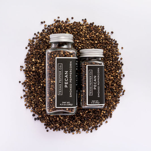 Two bottles of Texas Pepper Co. pecan smoked peppercorn surrounded by pepper corns on a white background.