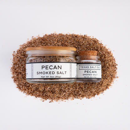 Top-down view of Texas Salt Co. Pecan Smoked Salt, showing 3 oz jar with bamboo lid and 0.5 oz jar with cork lid, placed on a bed of coarse pecan smoked salt.