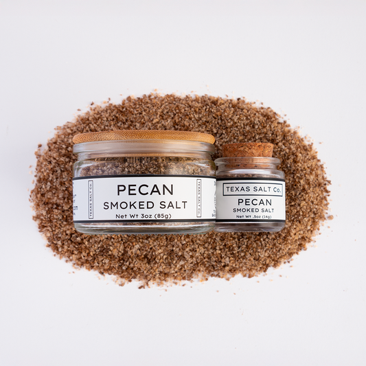 Top-down view of Texas Salt Co. Pecan Smoked Salt, showing 3 oz jar with bamboo lid and 0.5 oz jar with cork lid, placed on a bed of coarse pecan smoked salt.