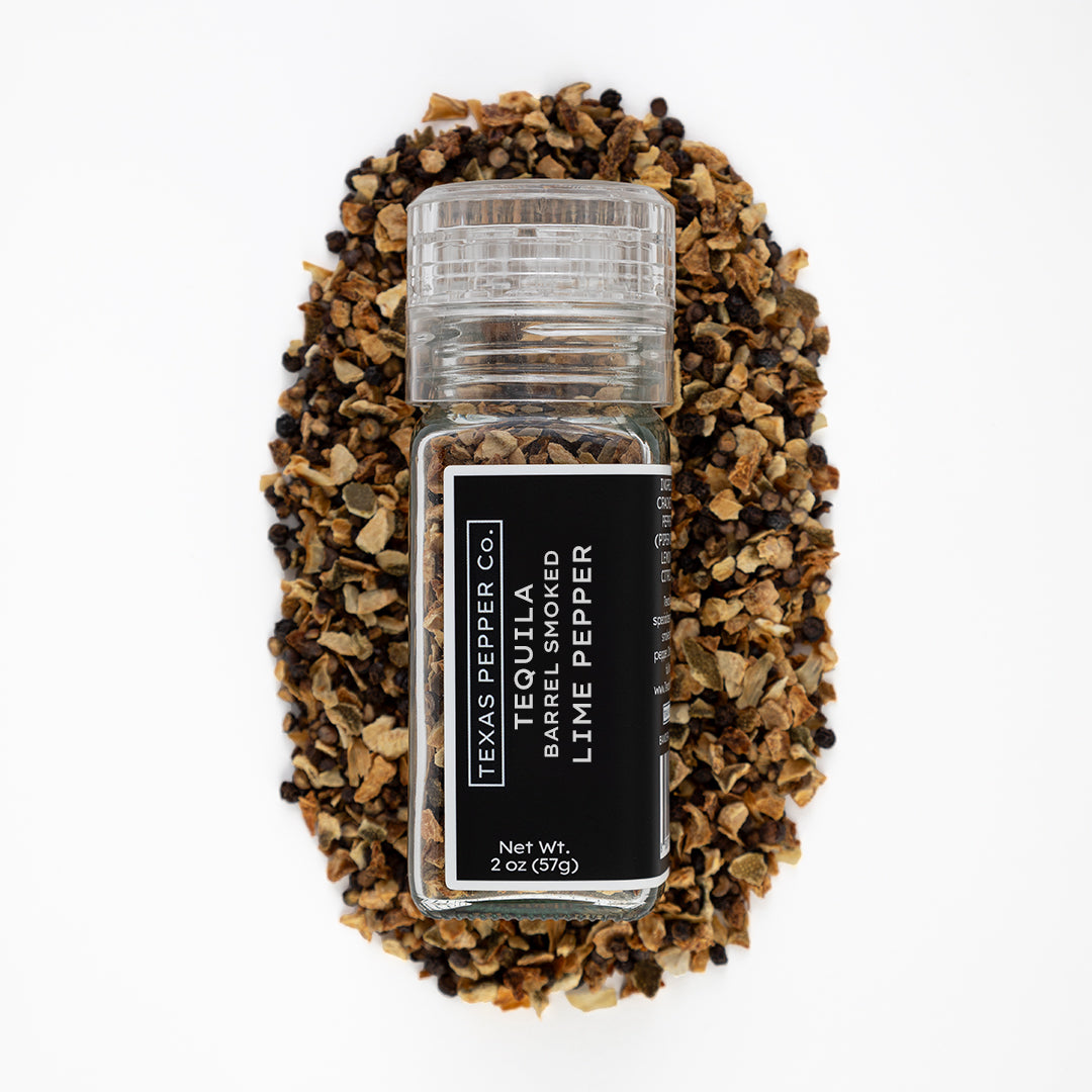 Spice container labeled 'Lime Pepper' surrounded by a pile of dried lime rinds and half cracked peppercorn on a white background